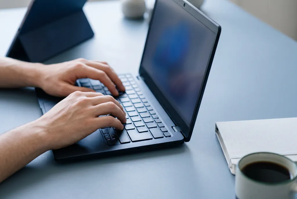 Hands typing on a laptop with the screen turned away, beside a notebook and a coffee.